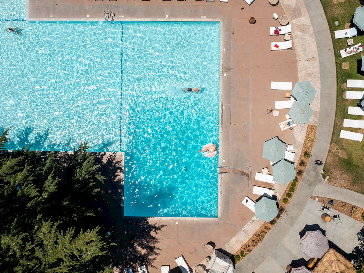 An aerial view of a swimming pool surrounded by lounge chairs and umbrellas, with a few people swimming and relaxing in the water.