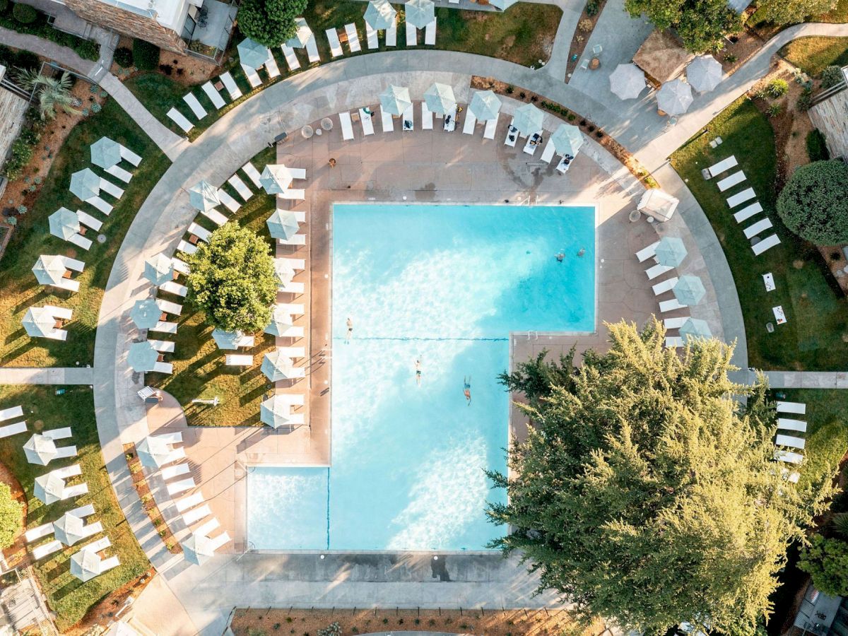 The image shows an aerial view of a large rectangular swimming pool surrounded by numerous lounge chairs and trees, within a landscaped area.