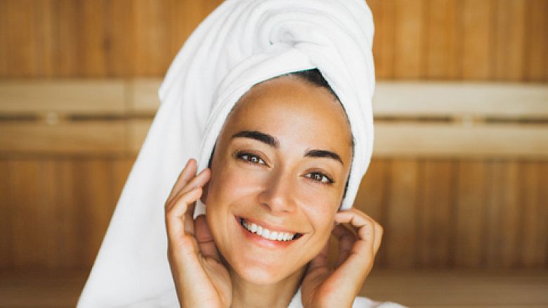 A person in a white bathrobe and towel, smiling in a wooden sauna room, hands near face.
