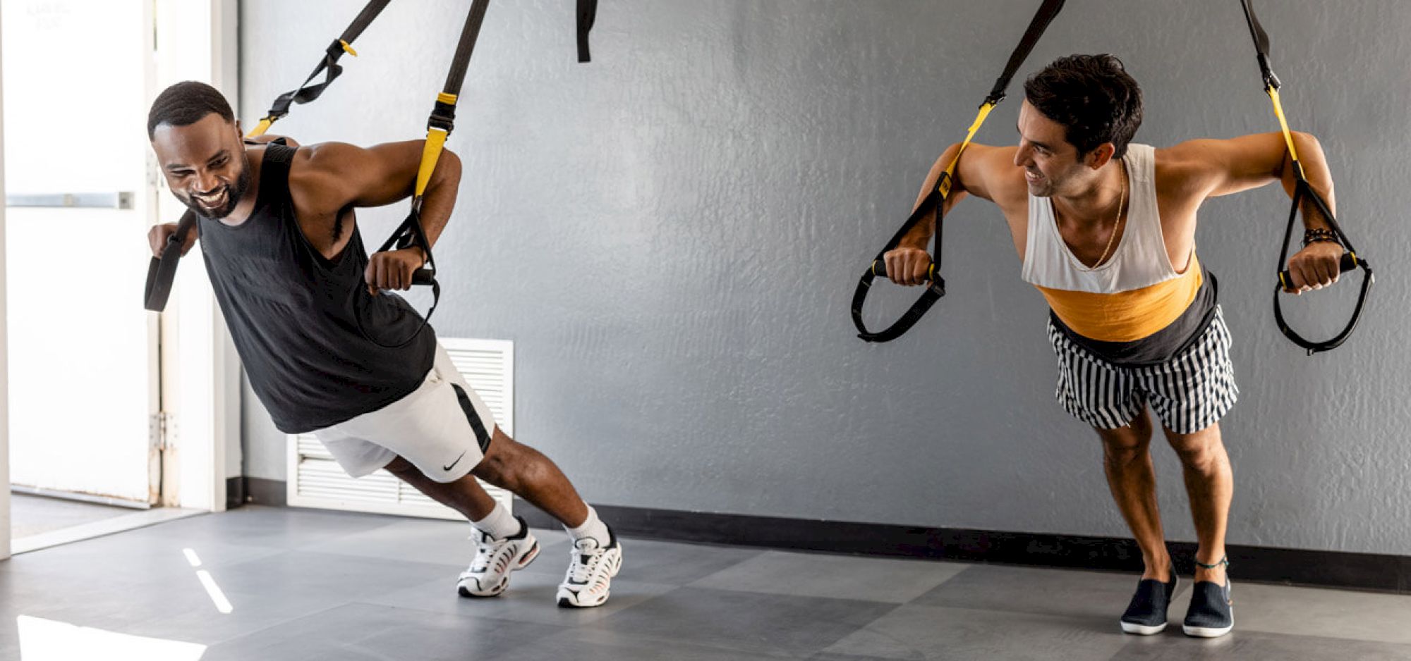 Two men are doing a suspension training exercise while smiling at each other in a gym. They are using TRX straps anchored to the ceiling.