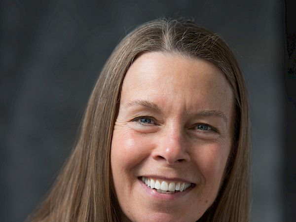 A smiling woman with straight, shoulder-length hair is wearing a dark shirt. She is standing against a dark, neutral background.