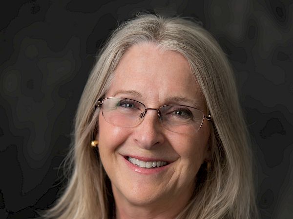 A person with long, blonde hair and glasses is smiling, wearing a black shirt with an embroidered logo. The background is dark.