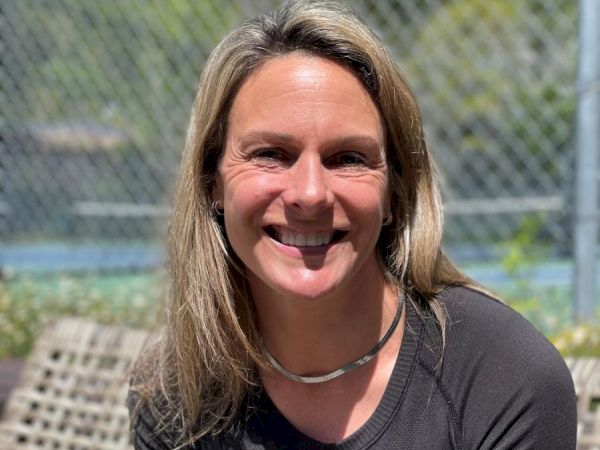 A person with long blonde hair is smiling and wearing a black top while sitting outside on a bench; there's a chain-link fence and greenery in the background.