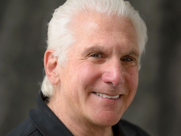 A man with white hair smiles at the camera, wearing a dark collared shirt with a subtle logo, set against a blurred background.