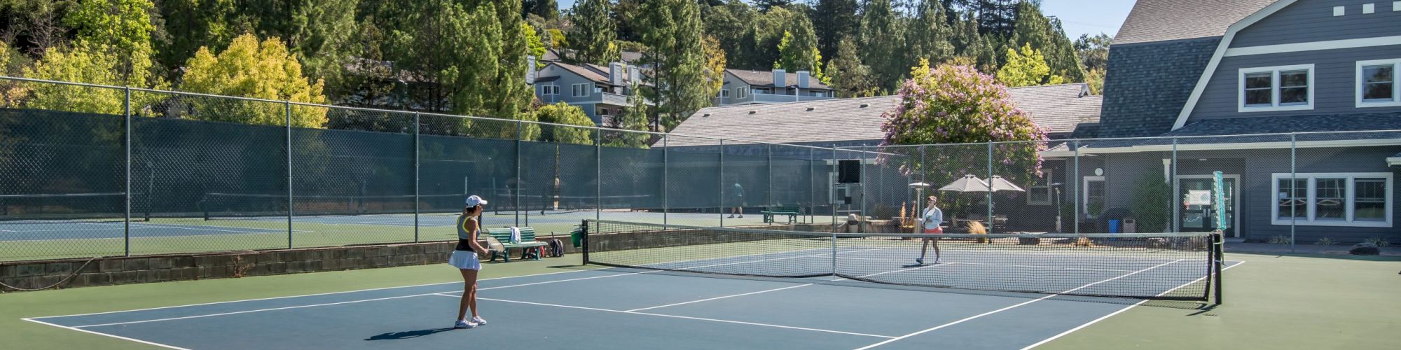 Two people are playing tennis on an outdoor court surrounded by trees and buildings.