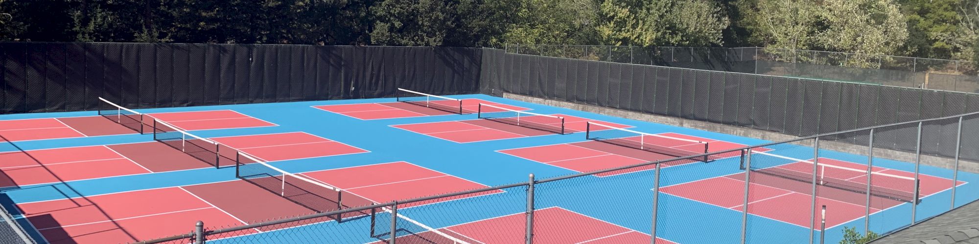 The image shows multiple pickleball courts with red surfaces and blue outlining, surrounded by fencing and trees in the background.