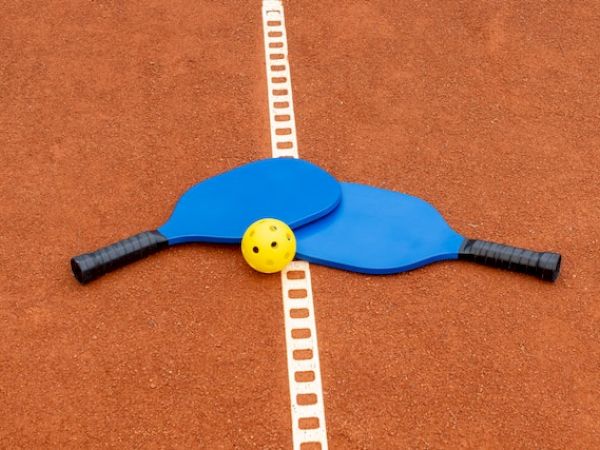 The image shows two blue pickleball paddles crossed over each other with a yellow perforated plastic ball placed on top, on a court.