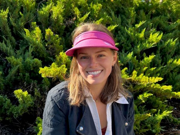 A person wearing a magenta visor and black jacket is smiling in front of lush green bushes and trees, enjoying a sunny day outdoors.