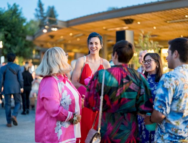 A group of people in colorful outfits are socializing outdoors at an event in the evening, under a well-lit, wooden-roofed area.
