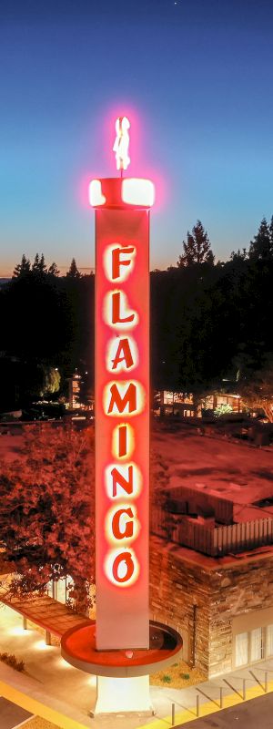 A tall, illuminated "FLAMINGO" sign stands over a building at dusk, surrounded by parking spaces and trees in the background.