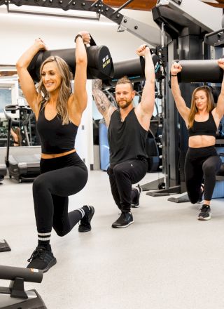 Three people are doing weighted lunges in a gym, lifting weights above their heads.