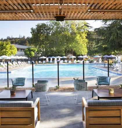 An outdoor seating area with couches and chairs overlooks a pool surrounded by lounge chairs and umbrellas, set amid greenery and trees.