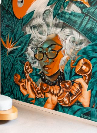 The image shows a stylish hotel lobby with a couple, luggage, modern furniture, and a large, colorful mural of a woman with white hair and glasses.