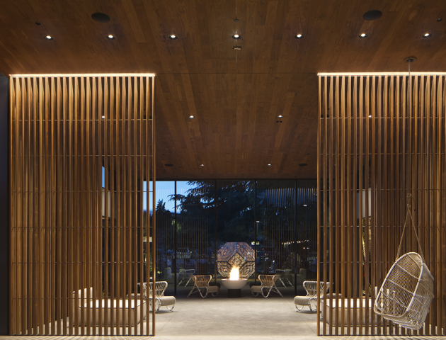 This image shows a modern outdoor patio area with wooden slats, hanging chairs, and a central seating space, well-lit and surrounded by trees.