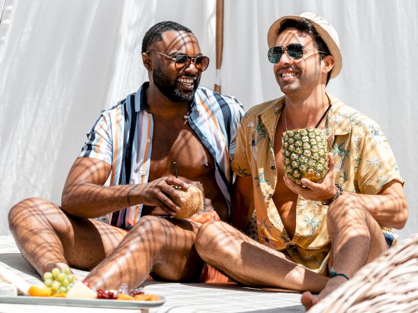 Two people are sitting and smiling in a sunlit, relaxed outdoor setting, each holding a drink, against a backdrop of white curtains.