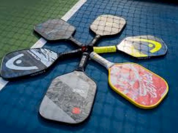 The image shows six pickleball paddles arranged in a star shape on a blue and green court surface.