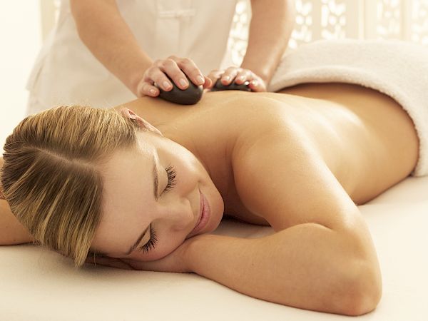 A person is lying on a massage table, receiving a hot stone massage on the back from a therapist, and appears to be relaxed.